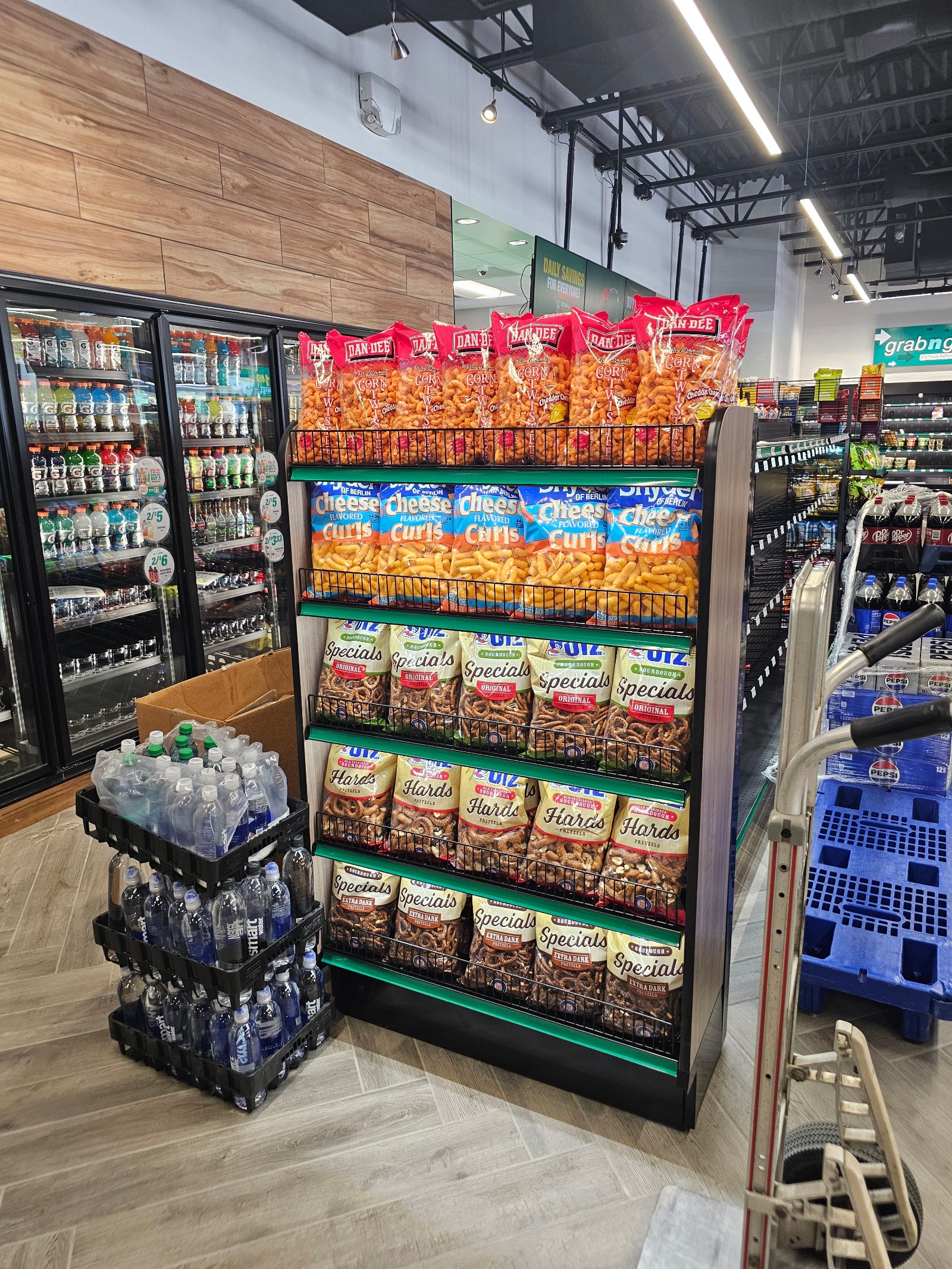 A merchandised convenience-store endcap caught mid-restock: an aluminum hand truck parked at the rack beside an open carton